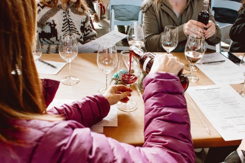 A person in a purple jacket pours red wine into a glass at a tasting event, surrounded by others and wine glasses.