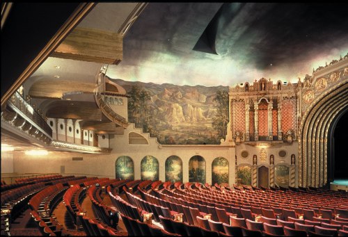 Interior of a vintage theater featuring ornate architecture, red seating, and a painted ceiling with scenic murals.