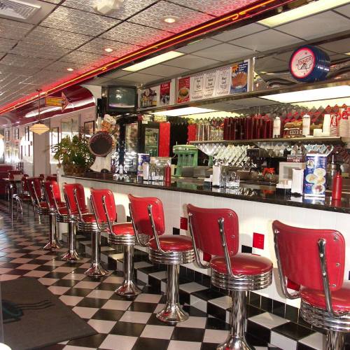 A retro diner interior featuring red stools, a checkered floor, and a long counter with vintage decor and menu signs.