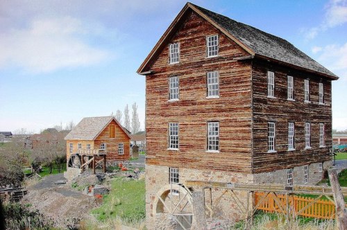 Two rustic wooden buildings, one tall with multiple windows and a waterwheel, set in a green landscape under a blue sky.