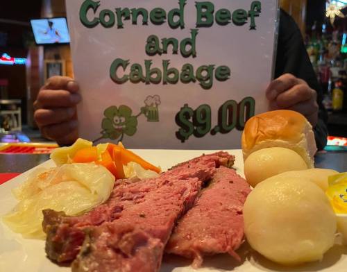 A person holds a sign reading "Corned Beef and Cabbage $9.00" next to a plate of corned beef, cabbage, and rolls.