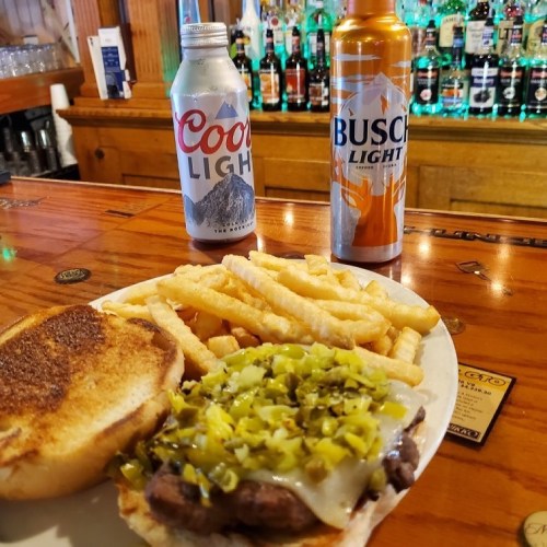 A plate with a cheeseburger topped with green peppers, served with fries, alongside two cans of beer on a bar counter.