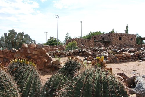 A desert landscape featuring stone ruins, cacti, and a clear sky with distant trees and light poles.