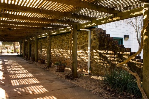 A sunlit walkway with wooden beams casting shadows on a stone wall, lined with potted plants and greenery.
