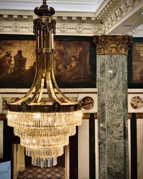 A grand chandelier hangs from a ceiling, surrounded by ornate columns and vintage artwork in a luxurious interior.