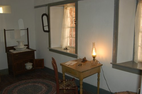 A simple room with a wooden desk, chair, oil lamp, and a washstand by the window, featuring natural light and rustic decor.