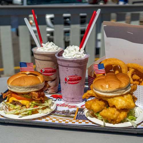 A tray with two burgers, onion rings, and three milkshakes topped with whipped cream and mini American flags.