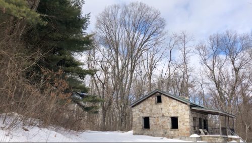 Abandoned stone house surrounded by snow-covered ground and bare trees under a cloudy sky.
