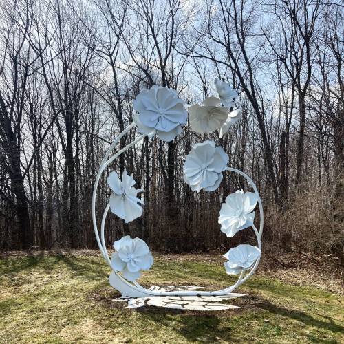 A white floral sculpture surrounded by trees, set in a grassy area under a clear blue sky.