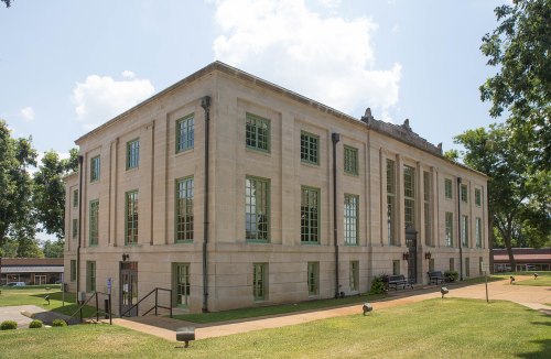 A large, stone building with green windows, surrounded by trees and a grassy area under a blue sky.