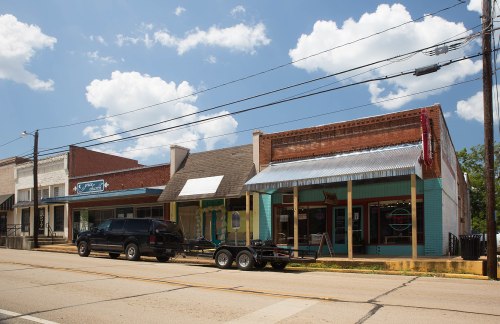 A small town street featuring several storefronts, a black truck with a trailer parked in front, under a blue sky.