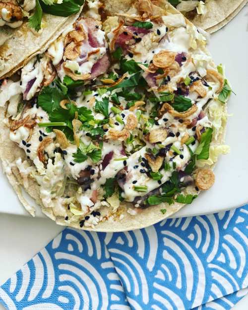 Close-up of tacos topped with shredded cabbage, herbs, crispy onions, and a creamy sauce on a patterned napkin.