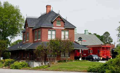 A historic brick house with a steep roof, next to a red train caboose and surrounded by greenery.