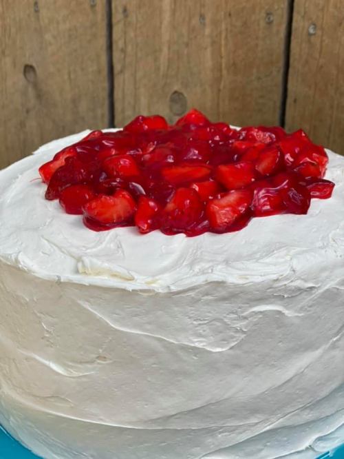 A round cake with white frosting topped with a layer of red jelly and fresh strawberries.