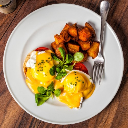 A plate of eggs Benedict with poached eggs, hollandaise sauce, tomatoes, and seasoned potato wedges.