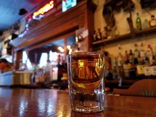 A close-up of a shot glass filled with amber liquid, set on a wooden bar with bottles in the background.