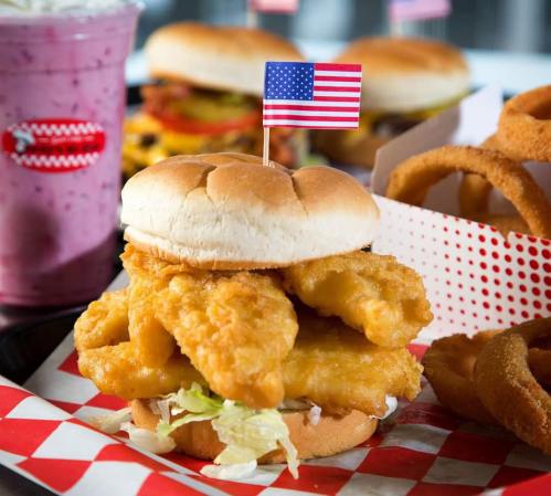 A crispy fish sandwich topped with lettuce and a small American flag, served with onion rings and a milkshake in the background.