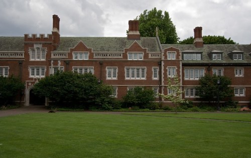 A large brick building with multiple windows and a manicured lawn, set against a cloudy sky.