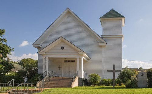 A white church building with a green roof, steps leading to the entrance, and a cross in the foreground.