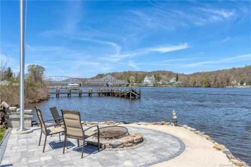 A serene riverside view with chairs around a fire pit, a dock in the distance, and trees lining the shore under a blue sky.