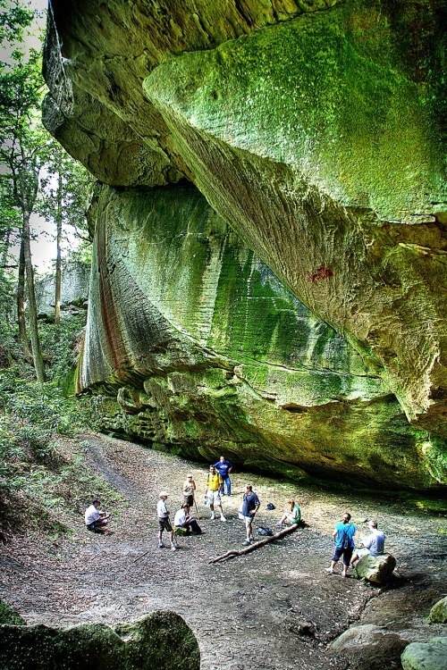 A group of people gathered under a large, moss-covered rock formation in a forested area.