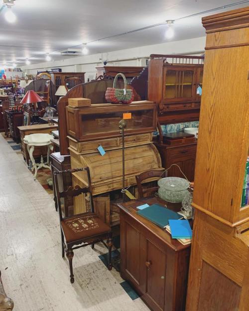 A variety of vintage furniture pieces displayed in a well-lit antique shop, including tables, cabinets, and lamps.