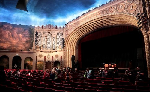 Interior of a grand theater with ornate architecture, red seats, and a stage set for a performance. Audience mingling.