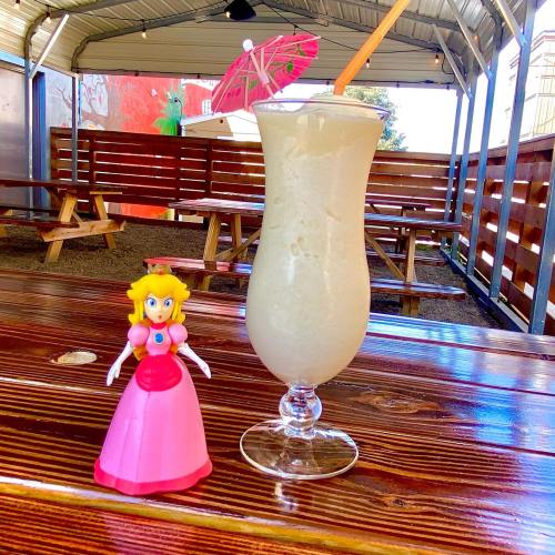 A pink princess figurine next to a tropical drink with a small umbrella, set on a wooden table in a casual outdoor space.