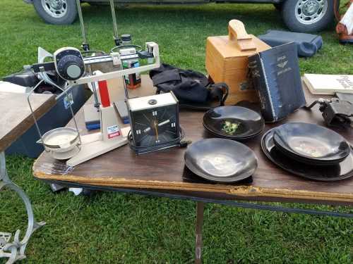 A table displays vintage items: a scale, clock, plates, and a wooden box, set on a grassy outdoor area.