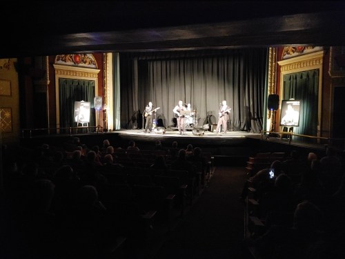 A band performs on stage in a theater, with an audience seated in the dimly lit background.