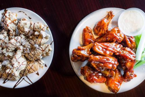 A plate of crispy chicken wings in sauce next to a plate of dessert drizzled with chocolate and powdered sugar.