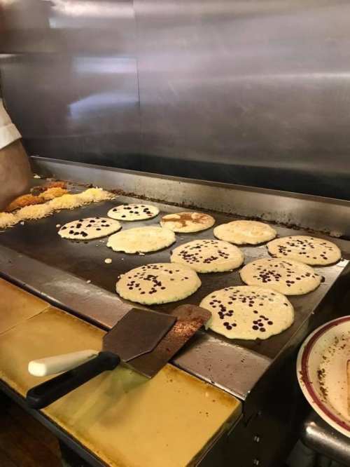 A griddle with several pancakes cooking, some topped with chocolate chips, in a busy kitchen setting.