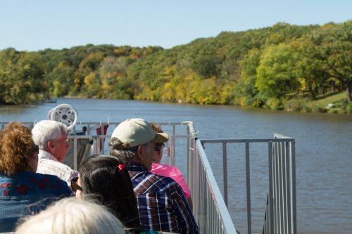 A group of people on a boat enjoying a scenic view of a river surrounded by trees in autumn colors.