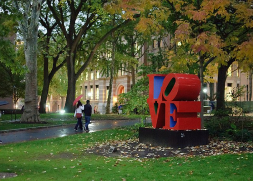 A colorful "LOVE" sculpture in a park with people walking under umbrellas amidst autumn foliage.