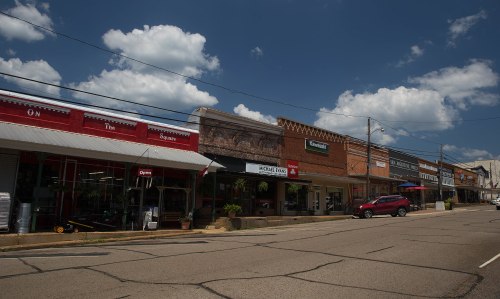 A small town street with brick storefronts, blue skies, and fluffy clouds, showcasing local businesses and a parked car.