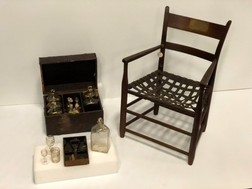 A wooden chair beside a vintage box containing glass bottles and small glasses on a white display platform.