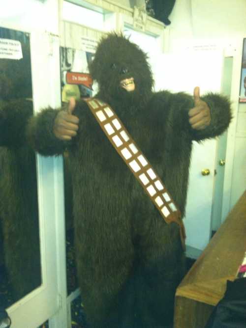 A person in a Chewbacca costume gives a thumbs up, standing in a room with mirrors and colorful decor.