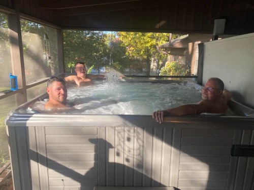 Three men relax in a hot tub, surrounded by a screened porch and greenery outside. Sunlight filters through the trees.