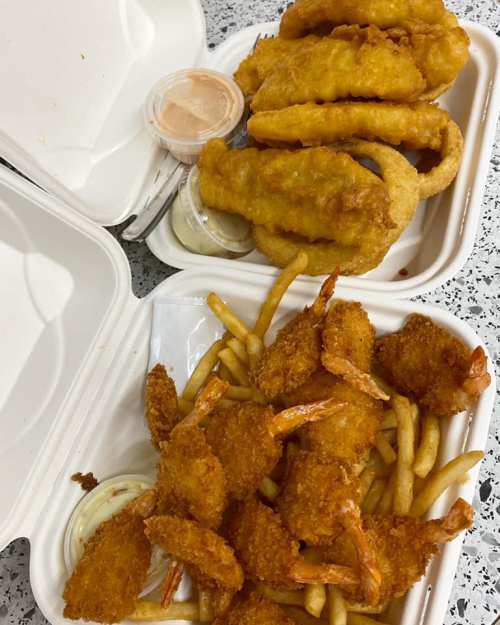 Two takeout containers filled with fried fish, shrimp, and fries, accompanied by dipping sauces.
