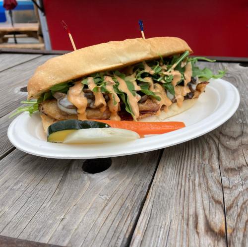 A large sandwich filled with grilled mushrooms, greens, and drizzled with sauce, served with pickles and carrots on a plate.