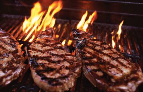 Grilled steaks with char marks sizzling over an open flame on a barbecue grill.