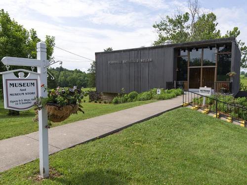 A modern museum building with a sign indicating it's open, surrounded by greenery and a pathway leading to the entrance.