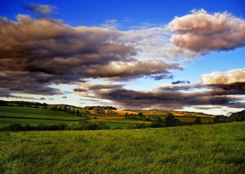 A vibrant landscape featuring green fields under a dramatic sky filled with fluffy clouds and a hint of sunset colors.