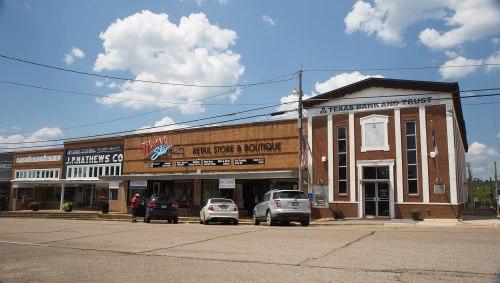 A street view of a small town featuring shops, a boutique, and a bank under a blue sky with fluffy clouds.
