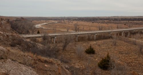 A winding road crosses a dry, open landscape with sparse trees and distant hills under a cloudy sky.
