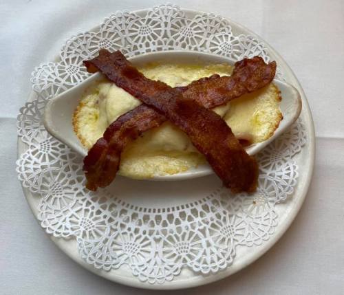 A creamy dish topped with melted cheese and two strips of crispy bacon, served on a decorative lace doily.
