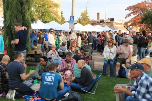 A lively outdoor event with crowds of people socializing, sitting on grass, and enjoying food under white tents.