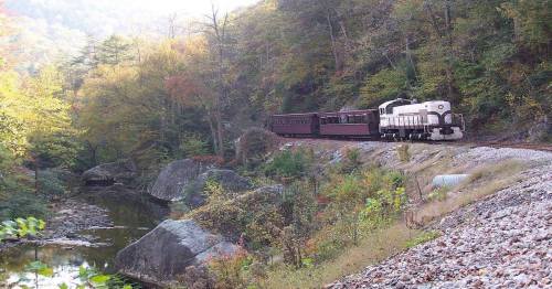 A vintage train travels along a scenic route beside a river, surrounded by colorful autumn foliage and rocky terrain.