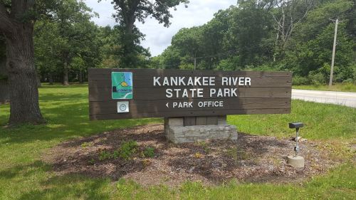 Sign for Kankakee River State Park, featuring a park office direction and a colorful logo. Green trees in the background.