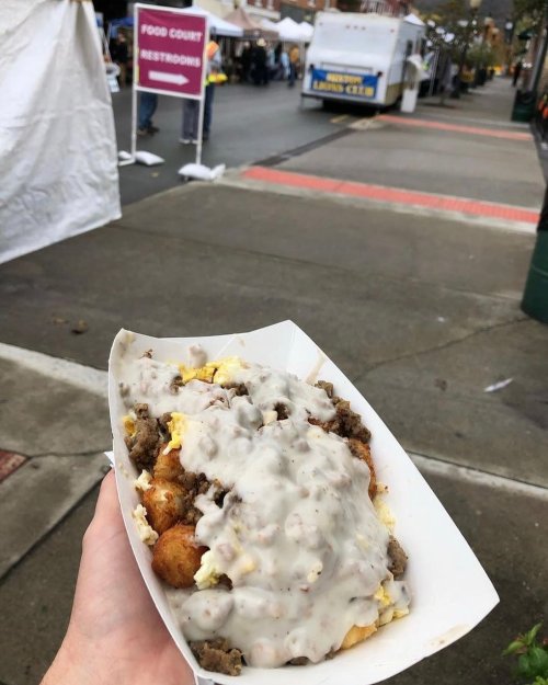 A hand holding a plate of loaded tater tots topped with sausage gravy, with food stalls in the background.